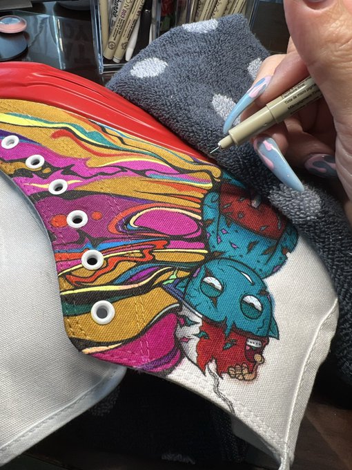 A close-up of a hand holding a fine-tipped marker, drawing intricate colorful artwork on a canvas shoe featuring a stylized teal skull character surrounded by pink, yellow, and red swirls.