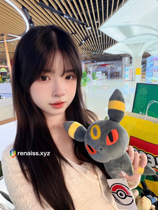 A selfie of a young woman holding a Umbreon Pokémon plush in what appears to be a mall, with the watermark 'renaiss.xyz' visible.