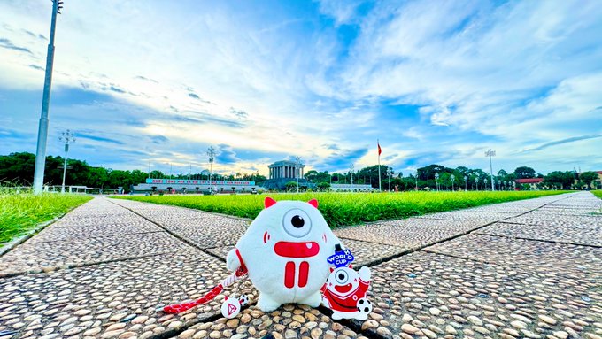 A plush toy mascot sits on a cobblestone path in Hanoi, Vietnam, with the Ho Chi Minh Mausoleum visible in the background under a blue sky.