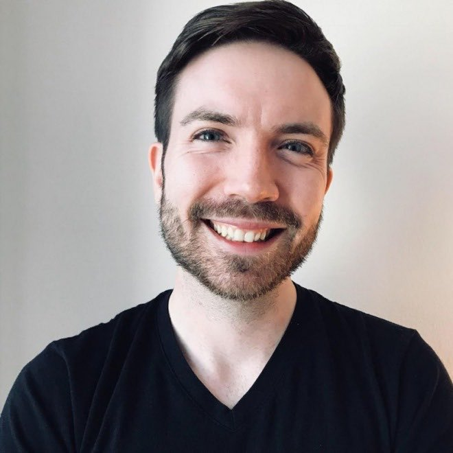 A portrait photograph of a smiling bearded man with dark hair wearing a black t-shirt against a plain light-colored background.