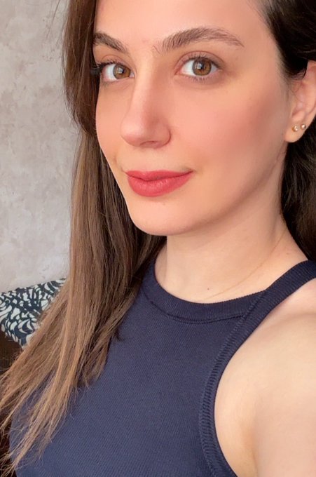 A close-up selfie of a young woman with brown hair, wearing a navy blue tank top and subtle makeup with pink lipstick.