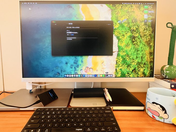 A desktop workspace setup features a monitor displaying a macOS login window over a tropical beach wallpaper, with a Rapoo wireless keyboard, a laptop, and a cartoon mug on the wooden desk.