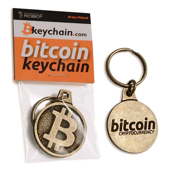 A brass-plated Bitcoin cryptocurrency keychain is shown both in its 'keychain.com' packaging and outside the bag.