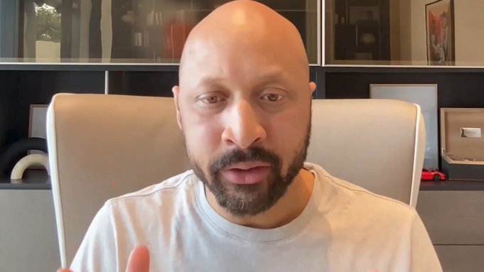 A bald man with a beard speaking directly to the camera from a chair in a modern office setting.