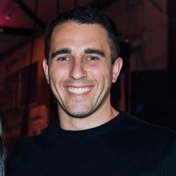 The image shows a smiling man with short dark hair wearing a black t-shirt, appearing to be at an event with a softly blurred background. This is likely Anthony Pompliano, the Bitcoin advocate referenced in the post, who is sharing his bullish outlook on BTC's price trajectory driven by AI profit rotation.