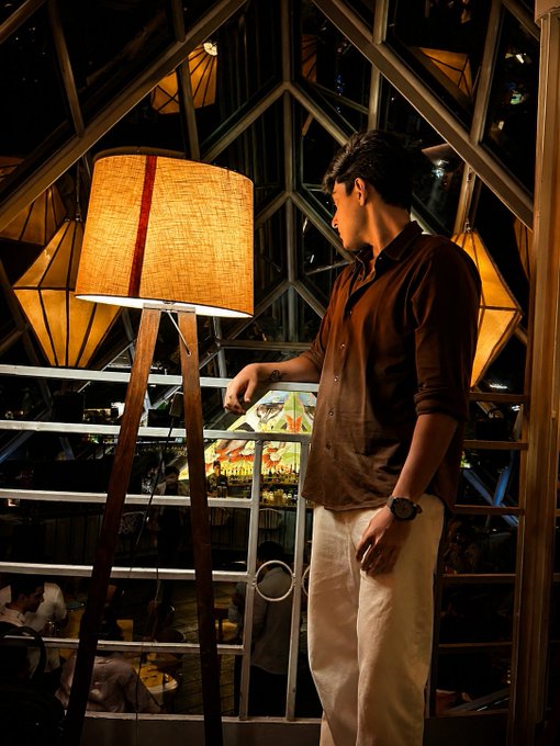 A young man in a brown shirt and light pants stands beside a large floor lamp with a fabric shade in a dimly lit venue with geometric window frames.