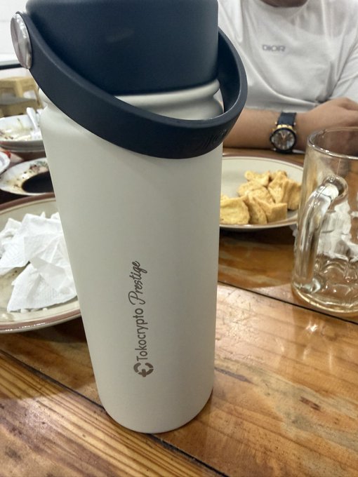 A white tumbler branded "Tokocrypto Prestige" sits on a wooden table, accompanying a meal of fried snacks and a clear glass mug, perfectly illustrating the caption about the tumbler being a loyal companion wherever the user goes.