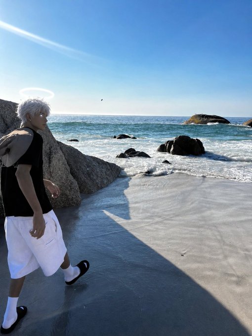 An anime-style or digitally edited figure with white hair, a halo, and white shorts walks along a beach shoreline with rocky outcrops and ocean waves under a clear blue sky.