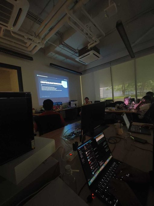 A dimly lit classroom or workshop setting where a presentation is being shown on a projector screen, with attendees working on laptops, including one displaying trading charts in the foreground.