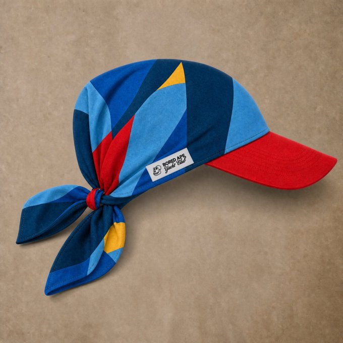 Product photo of a colorful bandana-style cycling cap with blue, red, and yellow geometric patterns, a red brim, and tied back, branded 'Bored Ape Yacht Club' on a small label.