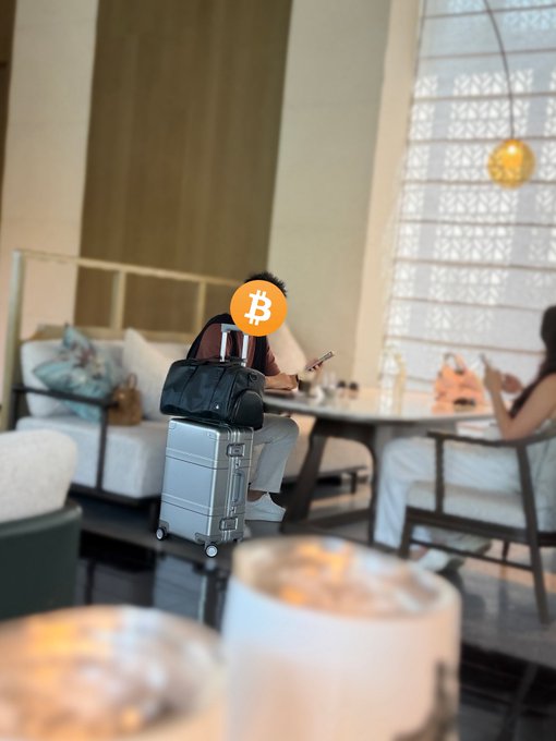 A person with a Bitcoin logo covering their face sits at a café table with a silver suitcase and black bag, while another person uses a phone nearby.