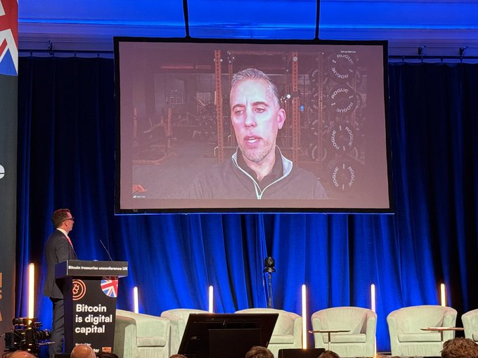 The image shows a man standing behind a podium, giving a speech or presentation at an event. The stage has a large screen displaying a video of the speaker talking about Bitcoin. There are several chairs arranged around the stage for attendees to sit and listen to the presentation. A banner is also visible in the background, possibly promoting the event or related topics.