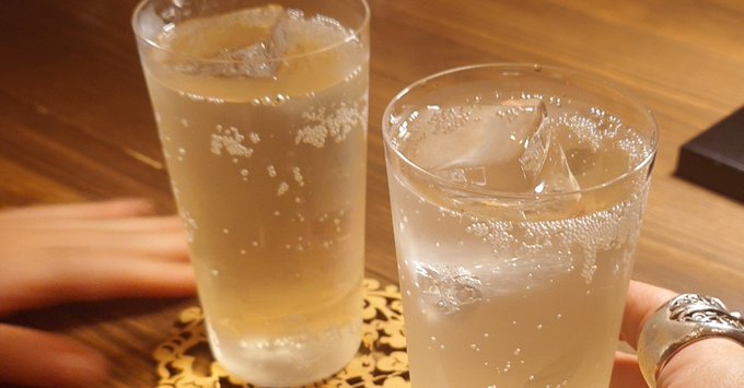 A close-up photograph shows two clear glasses filled with ice and a bubbly, light-colored beverage resting on a decorative coaster on a wooden table, with hands visible in the foreground.