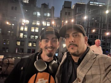 A photo of two smiling men posing together at night in a city setting with string lights overhead.