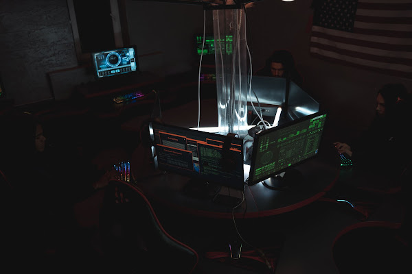 A dimly lit tactical operations room features multiple computer monitors displaying green text, with personnel working at stations around a central illuminated desk.