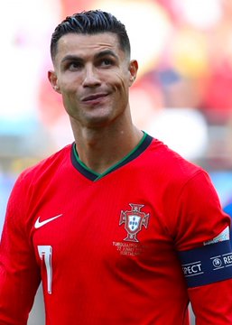 A photograph of soccer player Cristiano Ronaldo wearing a red Portugal national team jersey with the number 7 and captain's armband, smiling on the field.