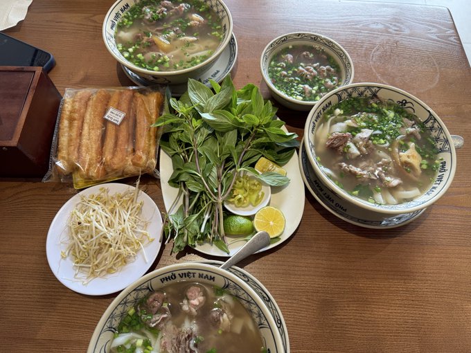 Three bowls of "Phở Việt Nam" noodle soup are arranged around a central plate of fresh herbs, lime, bean sprouts, and what appears to be chilis, with a side of breadsticks visible in the corner. The bowls contain rice noodles, broth, meat, and chopped scallions, complementing the post's description of a flavorful and reasonably priced Vietnamese meal in Ho Chi Minh City.