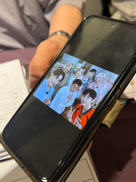 A hand holding a smartphone displaying a photo of a group of young people, including someone in an orange shirt, with what appears to be a receipt or printed paper visible underneath the phone.