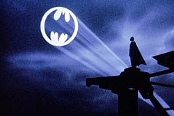A silhouette of Batman stands atop a building as the iconic Bat-Signal projects into the night sky, casting beams of light against the dark clouds. The glowing signal prominently displays the classic bat emblem, serving as a visual representation of the Tae Kim symbol referenced in the post.