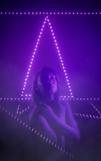 A woman stands with her eyes closed inside a room illuminated by purple LED lights arranged in a triangular pattern.