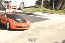 The image features a young child sitting inside an orange toy car that has "tgif" written on it. The child appears to be enjoying their time playing with the toy, possibly pretending they are driving or racing through the streets of a neighborhood. In the background, there is a white car parked next to a street sign and another car further away from the main scene.