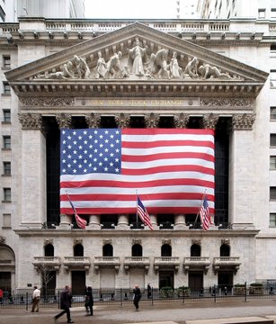 The New York Stock Exchange building is shown with a large American flag draped across its columned facade, with "NEW YORK STOCK EXCHANGE" visible in gold lettering on the pediment above. The patriotic imagery reinforces the post's announcement about NYSE bringing stock trading on-chain, symbolizing the integration of traditional American financial markets with cryptocurrency and blockchain technology.