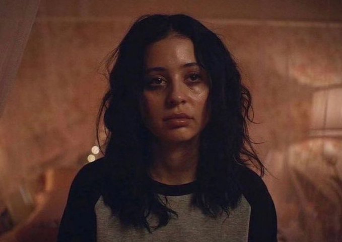 A close-up of a young woman with dark, wavy hair looking directly at the camera in a dimly lit, warm-toned setting.