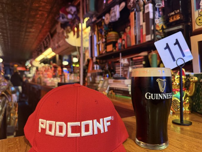 A red "PODCONF" cap sits on a wooden bar next to a pint of Guinness at PubKey, where table 11 marks the spot for a casual meetup blending Bitcoin talk, craft beer, and new connections.