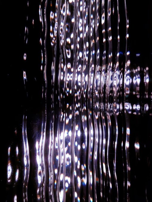 An abstract photograph showing vertical streaks and bokeh orbs of white, blue, and warm-colored lights reflected against a dark background, creating a distorted, shimmering visual effect.