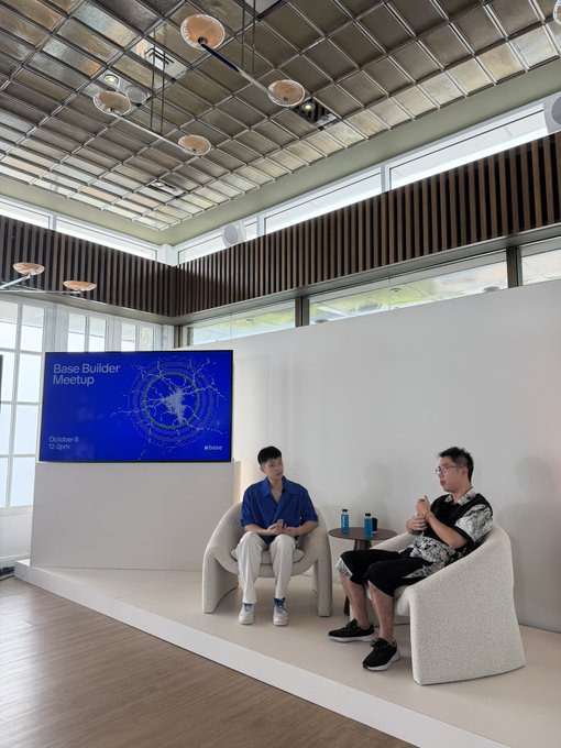Two men seated in white armchairs on a stage at a 'Base Builder Meetup' event (October 8, 11:30am), with a large screen displaying the event title and a world map graphic.