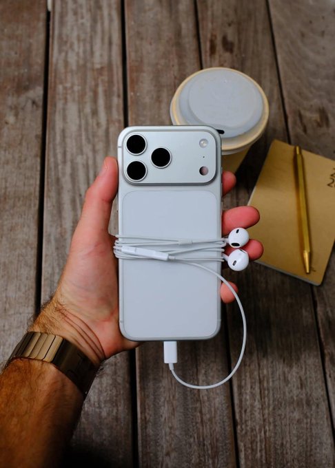 A hand holds an iPhone encased in a power bank cover with wired earbuds wrapped around it, set against a wooden plank background next to a coffee cup and notebook.