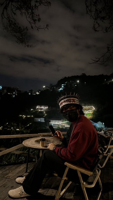 A person wearing a spiked crown, face mask, and dark red jacket sits on a wooden chair at night, looking at a phone with a scenic hillside view illuminated in the background.