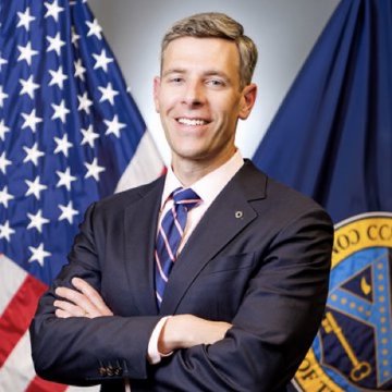 A professional portrait shows Jonathan Gould, OCC Comptroller, smiling with arms crossed in front of an American flag and what appears to be a Department of the Treasury seal. The image accompanies an announcement regarding his statement that final GENIUS Act stablecoin rules will be ready by November, with issuer applications potentially opening in 2027.
