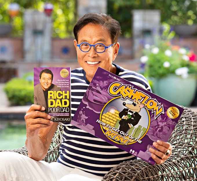 The image features a man sitting down with two books in his hands - one titled "Rich Dad Poor Dad" by Robert Kiyosaki and another named "Cashing In On The Rich Dad Poor Dad Game". He appears to be holding these books while posing for the camera, possibly discussing or promoting them.