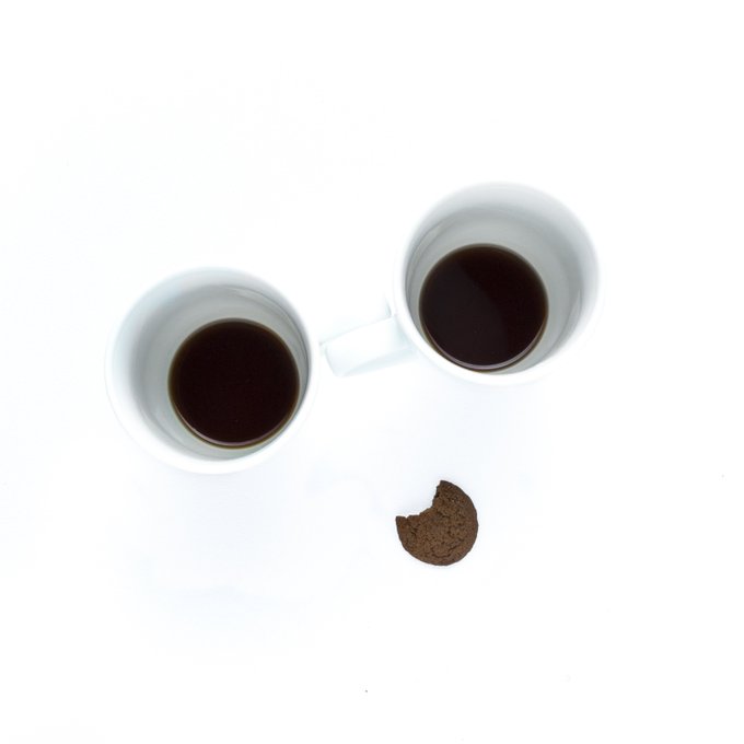 Two white mugs filled with black coffee are positioned facing each other on a white surface, with a single chocolate cookie featuring a bite taken out of it placed below them.