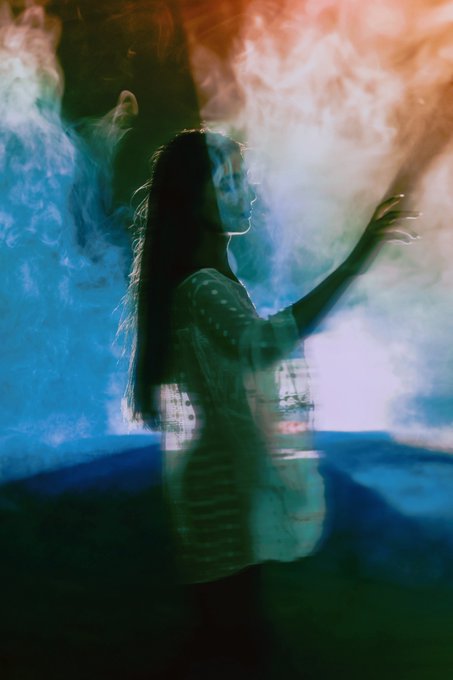 An artistic double-exposure photograph of a woman in profile with her hand extended, overlaid with hazy blue and orange light and smoke effects.