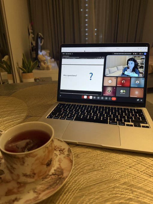 A laptop on a wooden table displays a video meeting with the text 'Any questions?', accompanied by a floral teacup filled with tea in the foreground.