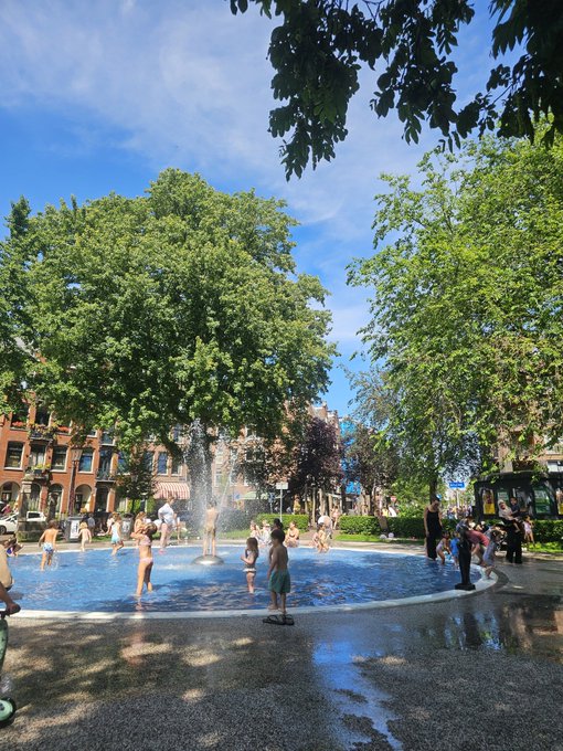 A sunny outdoor scene at a public splash pad/spray park, where children and adults are playing in the water fountains surrounded by large green trees and brick buildings.