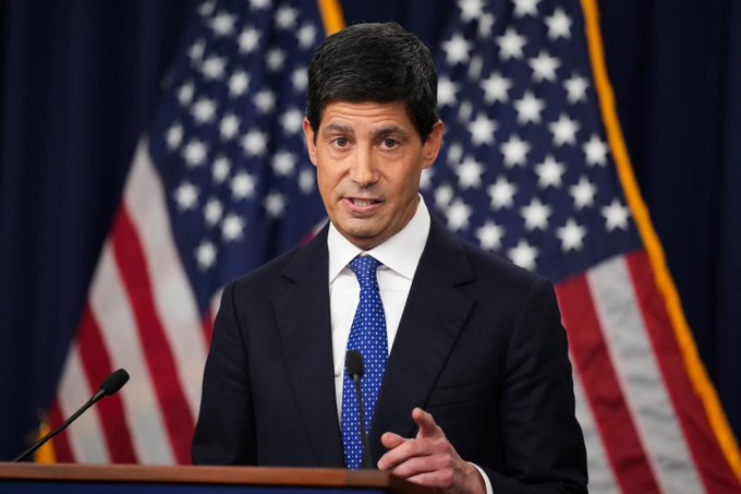 A man in a dark suit and blue tie speaks at a podium with two American flags behind him, appearing to be one of the eight Federal Reserve officials scheduled to deliver remarks during the macro-heavy week. His gesture and formal setting suggest an official monetary policy address.