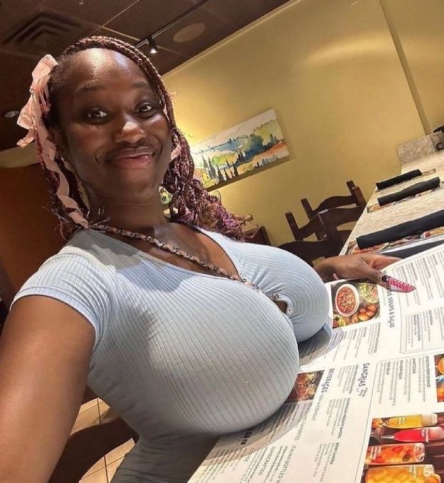 A woman with braided hair wearing a pink bow and a light blue ribbed top takes a selfie at a restaurant table covered with menus and paper placemats.