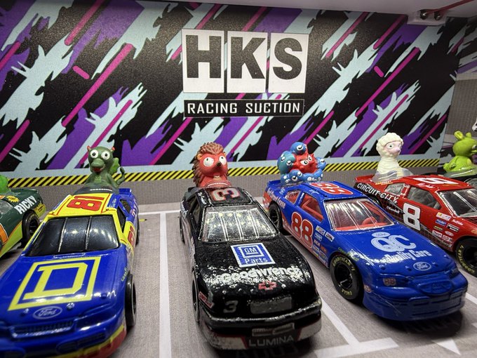 A display of NASCAR-style die-cast toy race cars (#3, #88, #8, etc.) is parked on a gray surface in front of a backdrop reading 'HKS RACING SUCTION' with various monster characters positioned as spectators.