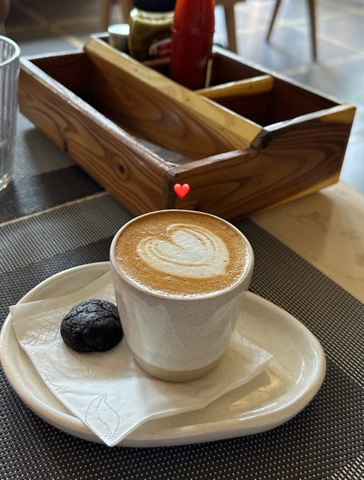 A cappuccino with heart-shaped latte art served in a speckled white cup on a saucer with a napkin and a small chocolate cookie, marked with a red heart emoji, on a restaurant table.