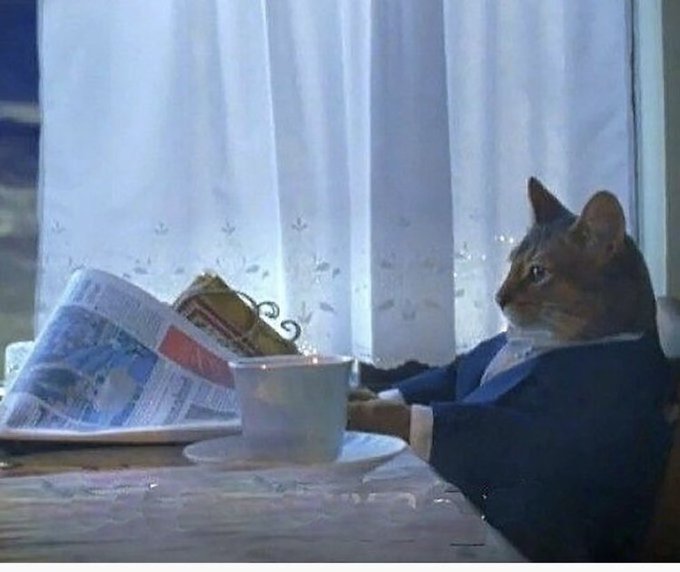 A cat dressed in a suit sitting at a table, appearing to read a newspaper while a teacup and saucer sit in front of it, with a lace curtain in the background.