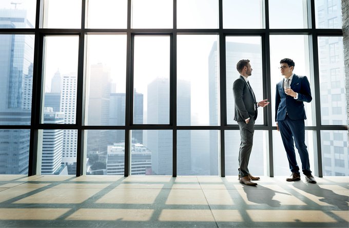 Two businessmen in suits converse by floor-to-ceiling windows overlooking a city skyline, visually reinforcing the post's theme of cross-border banking collaboration through platforms like Cosmos and Swift. The professional setting symbolizes the institutional partnerships enabling 24/7 global payments connectivity.
