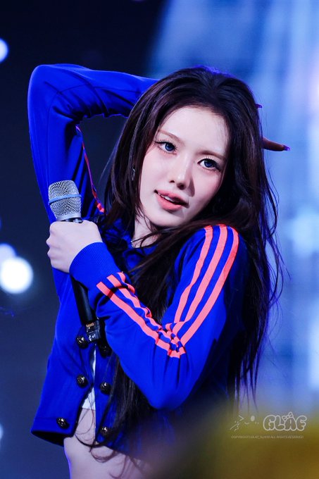 A young woman with long dark hair wears a blue Adidas jacket with orange stripes while holding a microphone on stage, striking a pose with her arms raised behind her head.