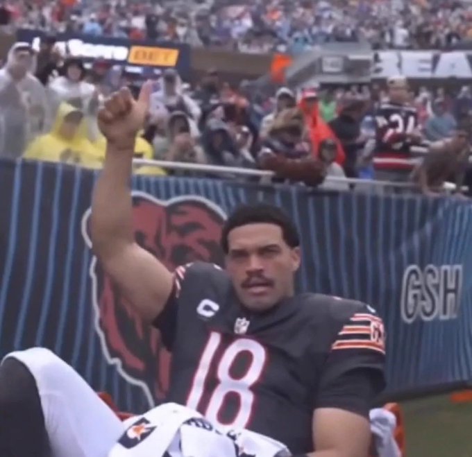 A Chicago Bears football player wearing jersey number 18 (Caleb Williams) sits on the field holding up one finger, with the GSH logo visible on the barrier behind him.