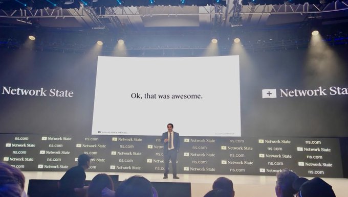 A man stands on a stage at a Network State conference, with a large screen behind him displaying the text 'Ok, that was awesome.'