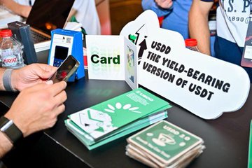 The image features a table with various items such as a cell phone, a sign, and some books. The main focus of the scene is a person using their cell phone while standing near these objects. There are also other people present in the background who might be interested in what's happening at this particular booth or event.