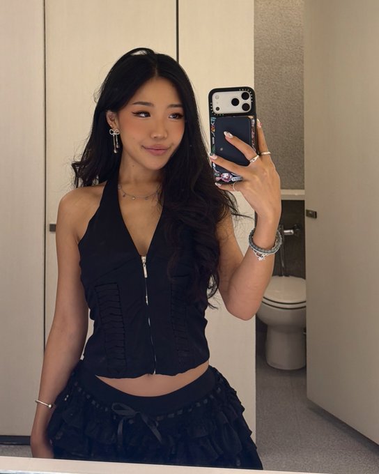 A young woman taking a mirror selfie in a bathroom, wearing a black halter top and skirt, holding a phone with a decorative case.