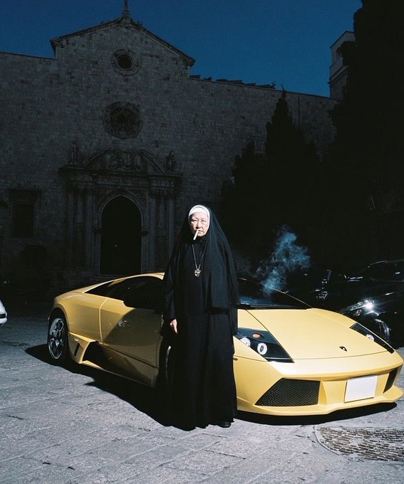 A striking photo of a nun smoking a cigarette while standing in front of a yellow Lamborghini Murciélago, with an old stone church in the background at dusk.